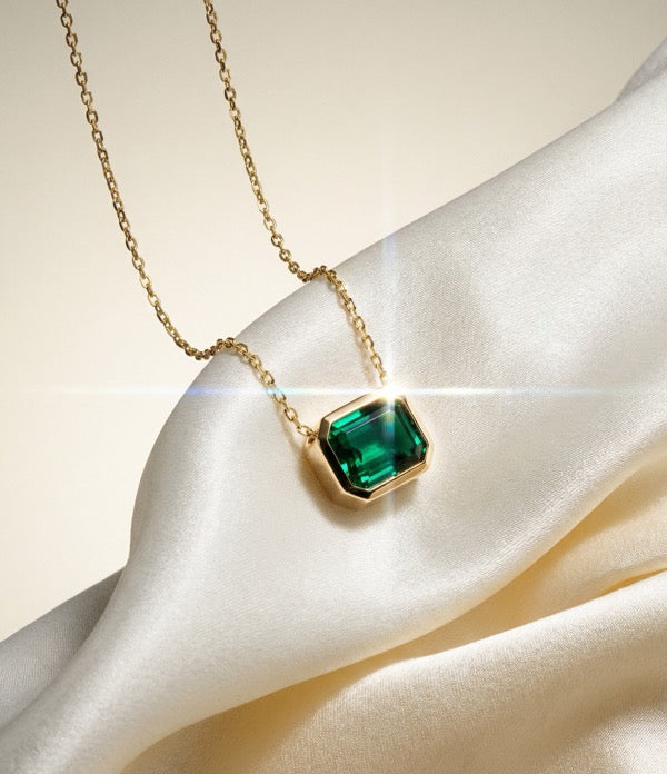 A green emerald-cut stone in a yellow gold bezel pendant on a fine chain, resting on ivory satin.