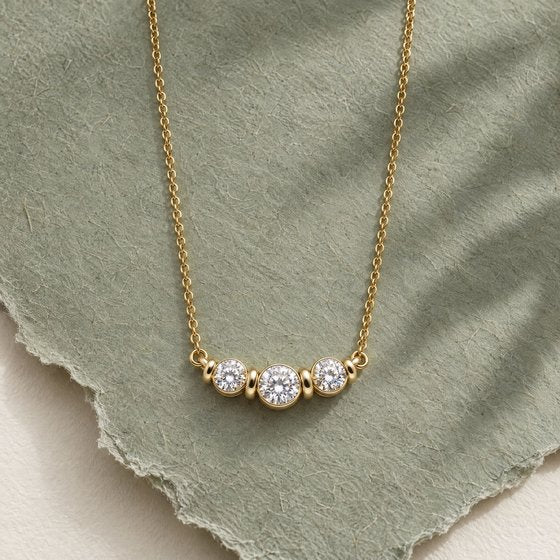A three-stone diamond bezel necklace in yellow gold laid on torn sage-green paper with a soft leaf shadow.