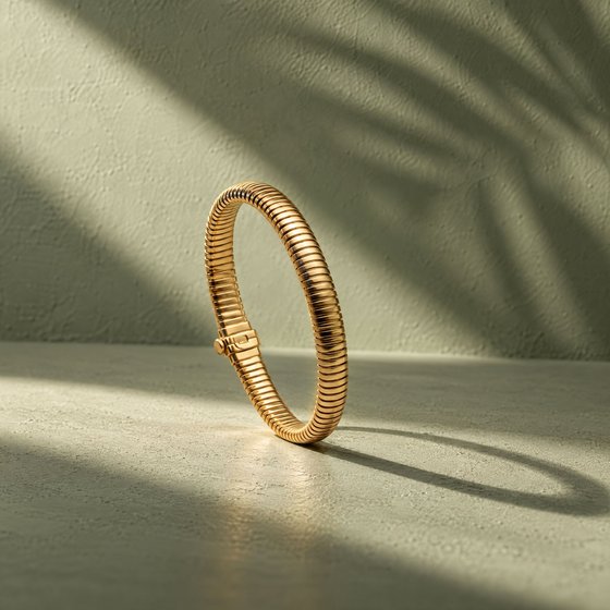 A slim ribbed gold tubogas bangle standing on sage plaster in a band of sunlight with a palm-leaf shadow.