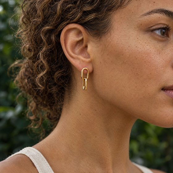 Close-up of a woman's ear wearing a delicate drop earring of two interlocking polished gold links, garden greenery behind.
