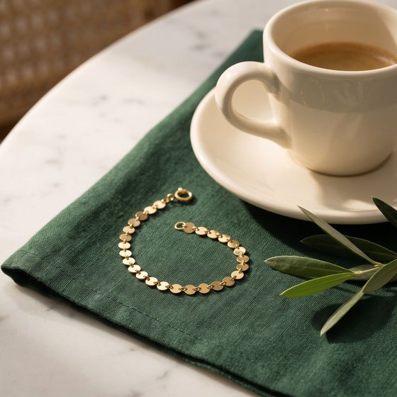 A delicate gold disc bracelet on a forest-green linen napkin beside an espresso cup and an olive sprig.