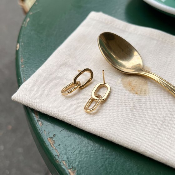 A pair of gold interlocking link drop earrings set down on an ivory linen napkin on a forest-green cafe table beside a teaspoon.