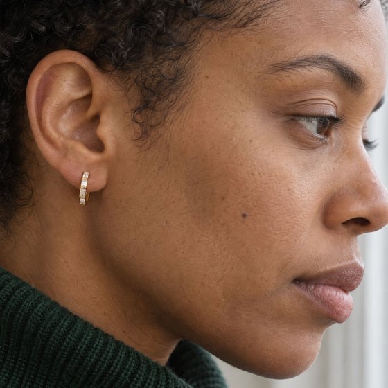 A woman in a forest-green turtleneck looking out of a window, wearing a small gold huggie set with baguette diamonds.