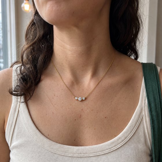 A woman's neckline in a white tank top, wearing a three-stone diamond bezel necklace in yellow gold.
