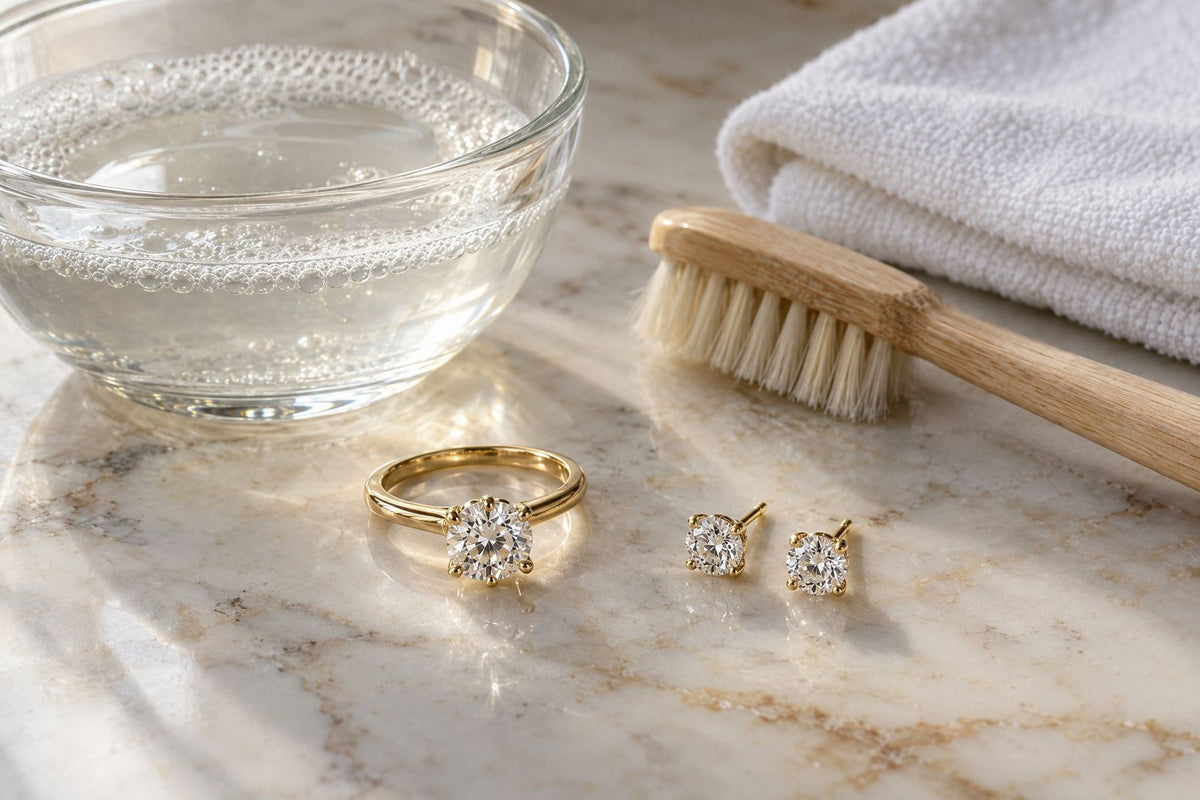 A diamond solitaire ring and diamond stud earrings beside a bowl of warm soapy water, a soft brush and a white cloth on cream marble.