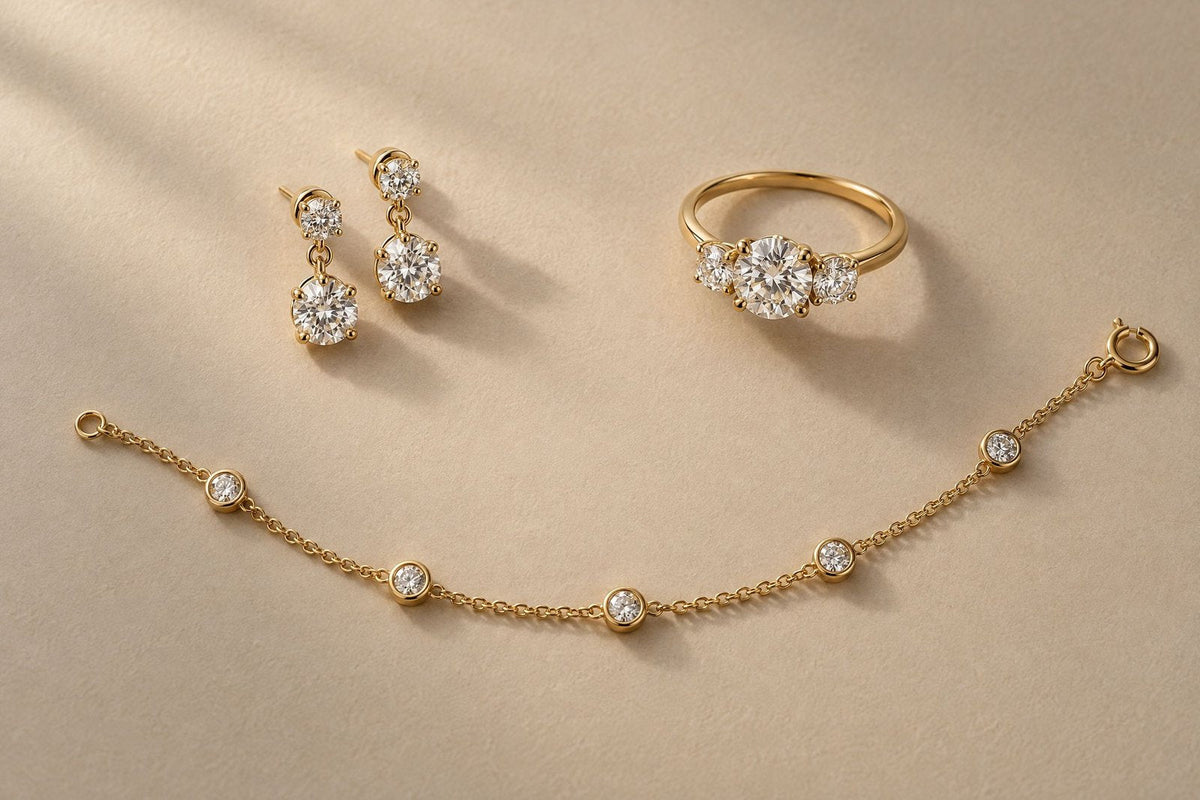 Diamond drop earrings, a diamond station bracelet and a three-stone engagement ring in yellow gold on warm sand paper.