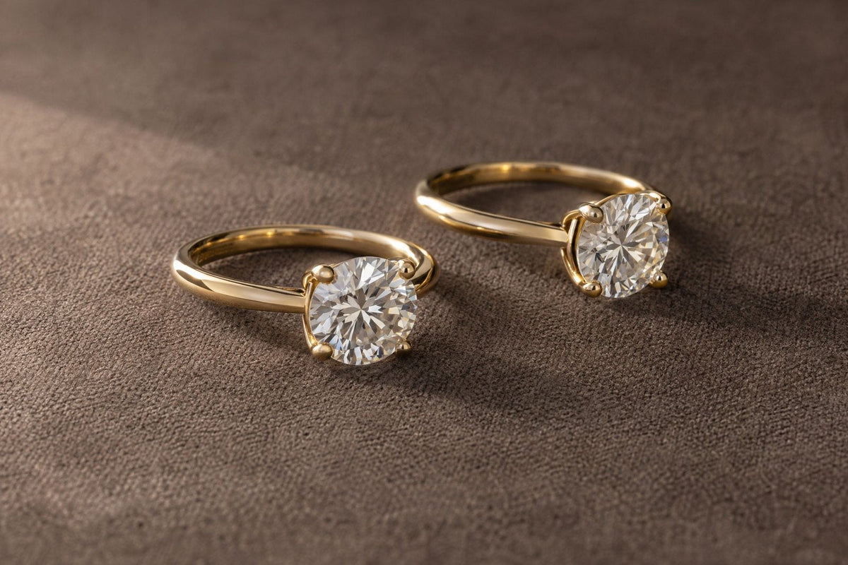 Two round diamond solitaire rings in yellow gold lying on their sides on taupe suede.