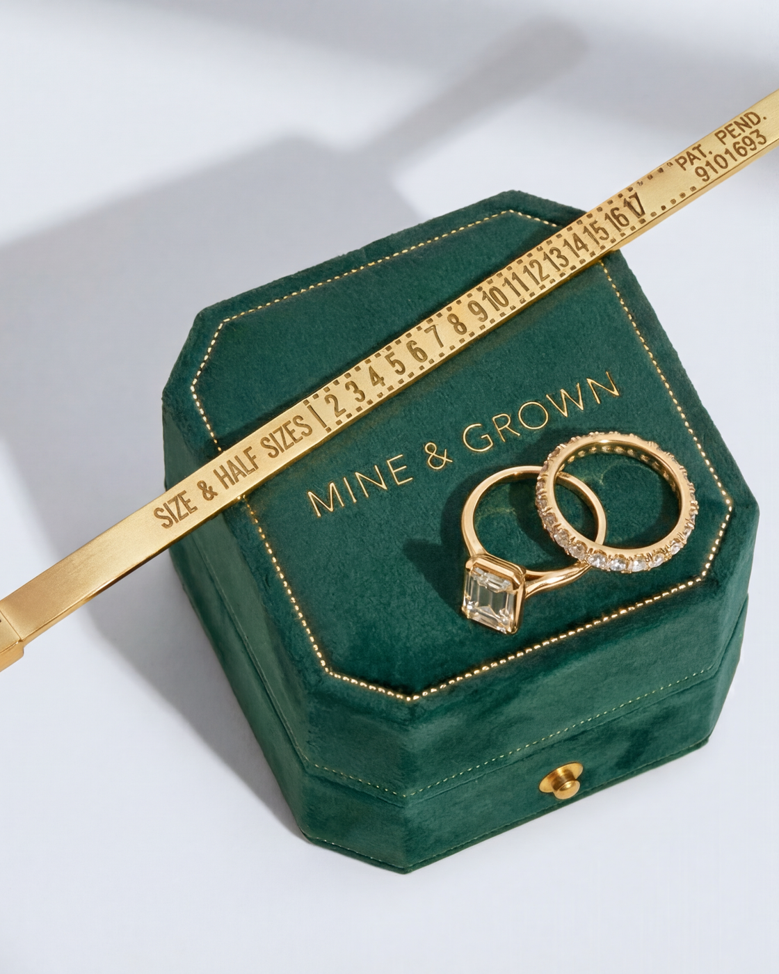 An emerald-cut engagement ring and a diamond band on a green velvet Mine & Grown ring box, beside a gold ring sizer