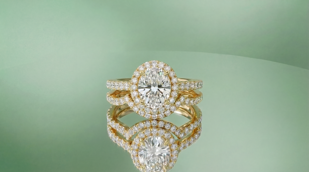 A yellow gold oval halo engagement ring stacked with a curved pavé band, reflected on a pale green surface