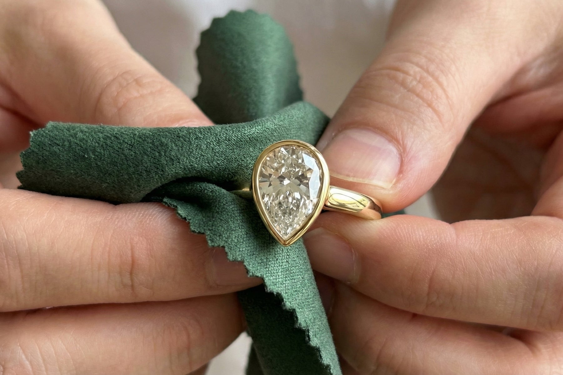 Hands polishing a pear-shaped diamond ring in a yellow gold bezel with a green cloth