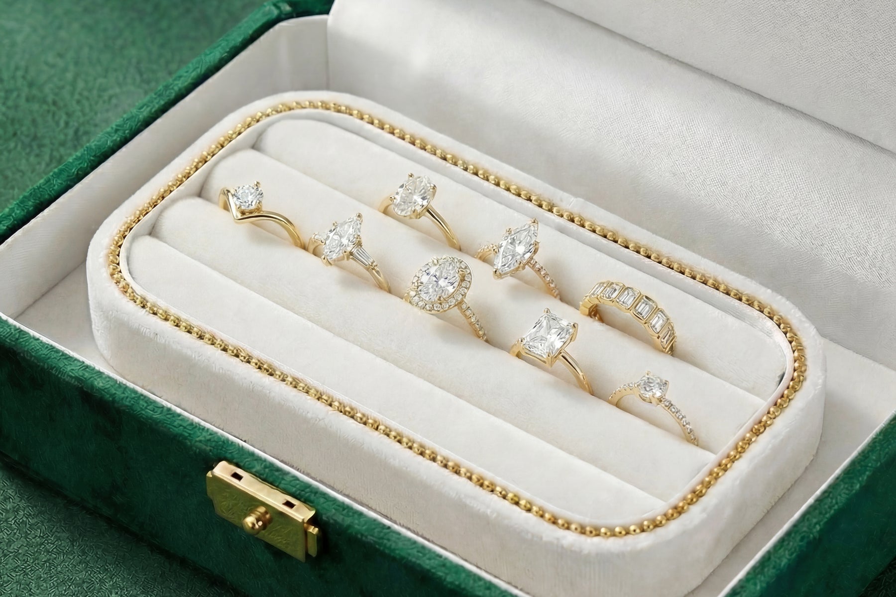 A green velvet jewelry box holding eight gold engagement rings with diamonds in different shapes