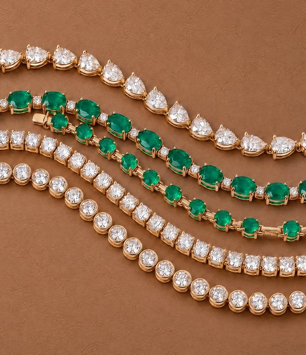 Five gold tennis bracelets laid in a gentle wave, set with pear and round diamonds and green stones.