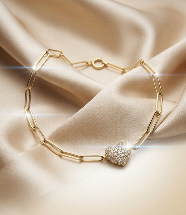 A yellow gold paperclip chain bracelet with a pavé diamond heart charm, on ivory satin.