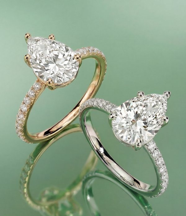 Two pear-shaped diamond engagement rings with pavé bands, one in yellow gold and one in white metal, on a sage green surface.