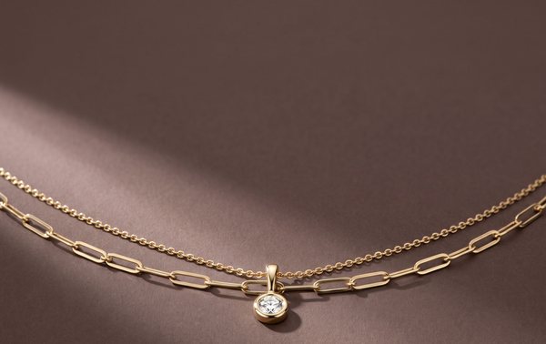 Two layered yellow gold necklaces, a paperclip chain and a cable chain with a bezel-set diamond pendant, on mocha paper.