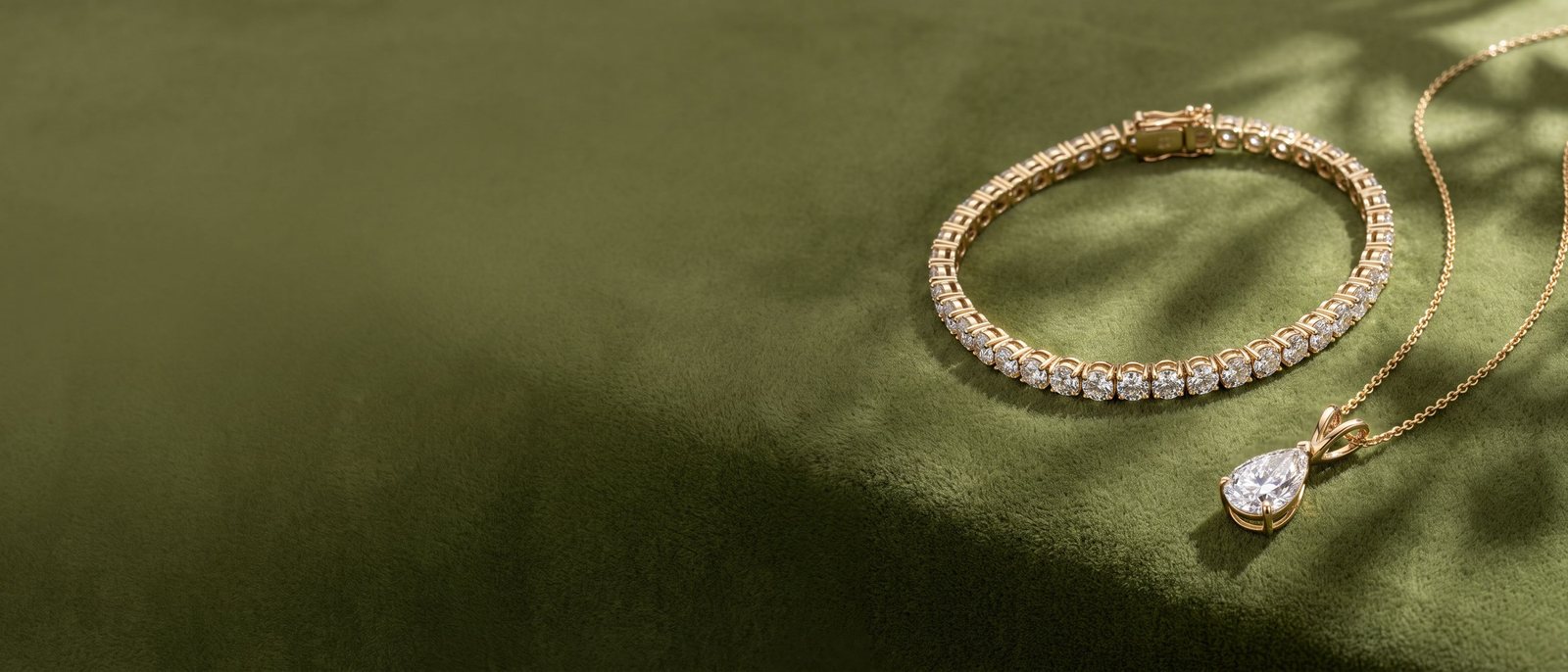 A diamond tennis bracelet and a pear diamond pendant in yellow gold on olive velvet with soft leaf shadows.