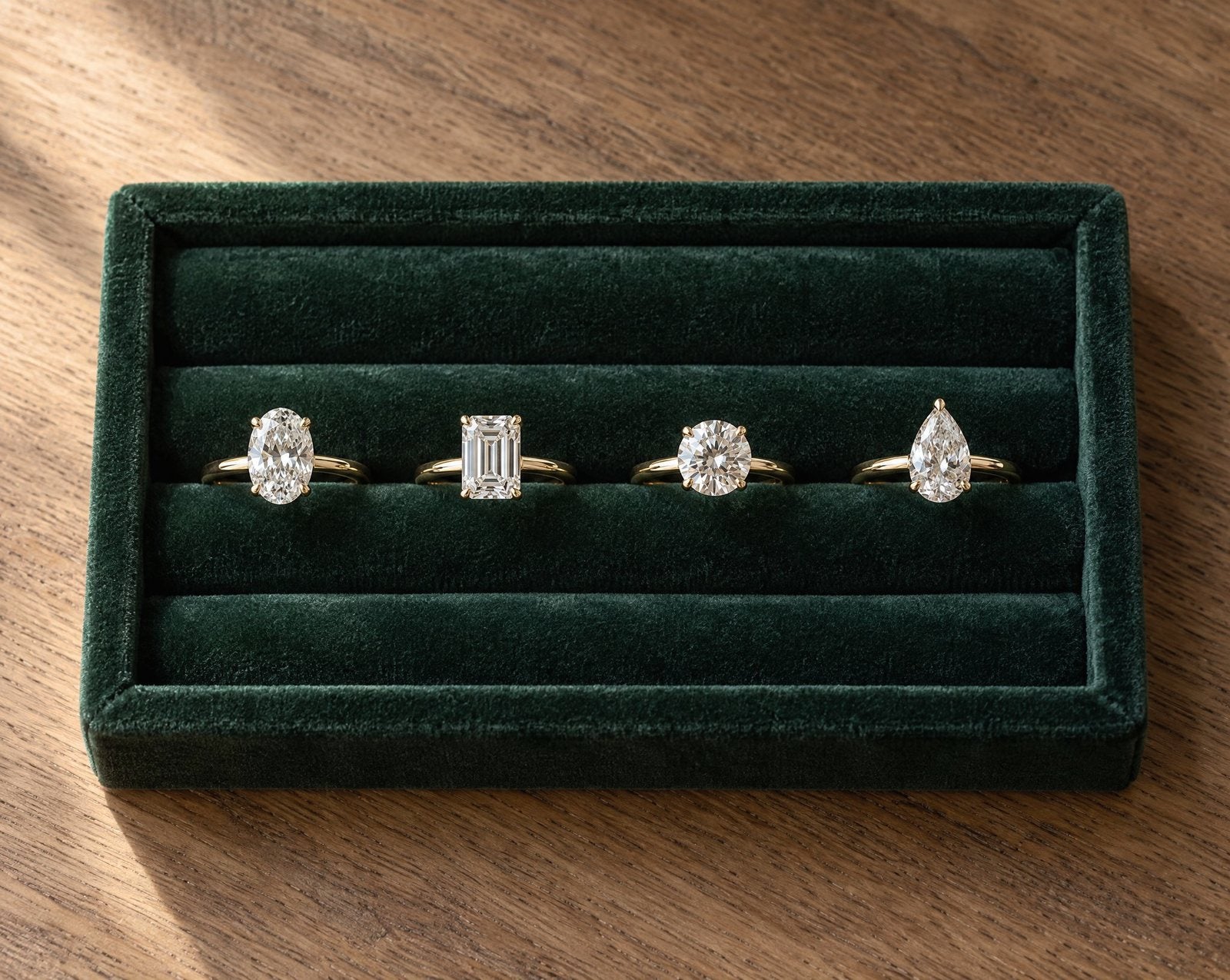 Four diamond engagement rings, oval, emerald, round and pear, in a green velvet ring tray on an oak table.
