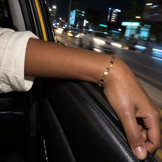A forearm resting on a taxi window at night, wearing a gold bracelet with small medallion charms.