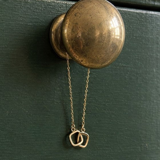 A gold necklace with two interlocking soft-square rings hanging from an antique brass knob on a forest-green painted dresser.