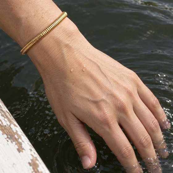 A woman trailing her fingers in lake water over the side of a boat, wearing a slim ribbed gold bracelet.