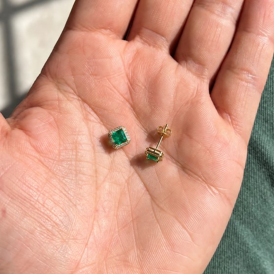 Two square emerald and diamond halo stud earrings in yellow gold resting loose in an open palm.