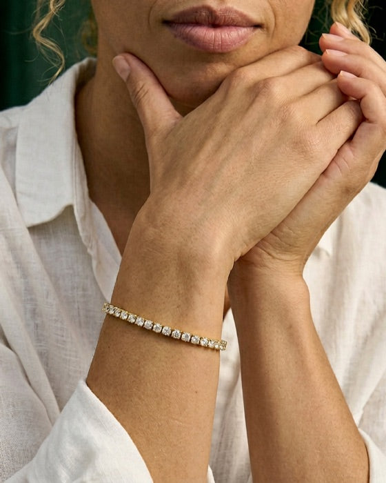 Hands raised to the chin wearing a gold tennis bracelet of round diamonds, against a green velvet curtain.