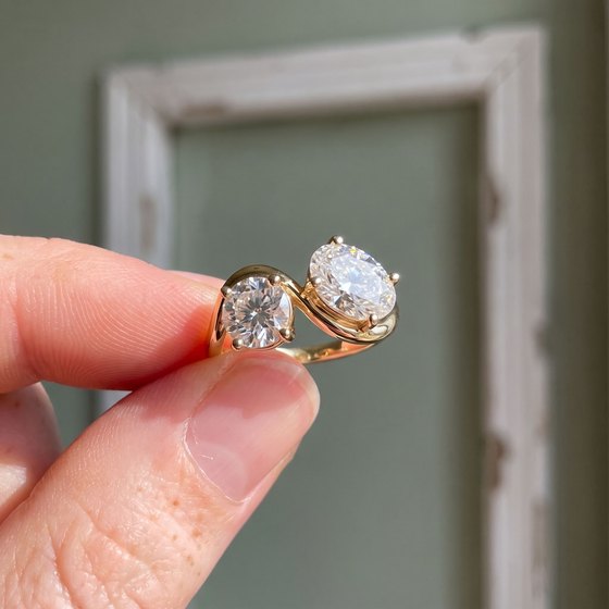 Fingertips holding up a yellow gold oval diamond ring against a sage-green wall.