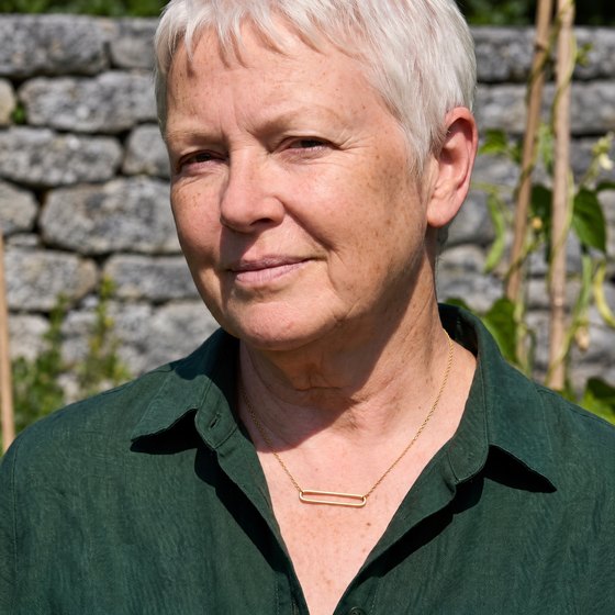 A white-haired woman in a forest-green linen shirt wearing an open rectangle gold link necklace.
